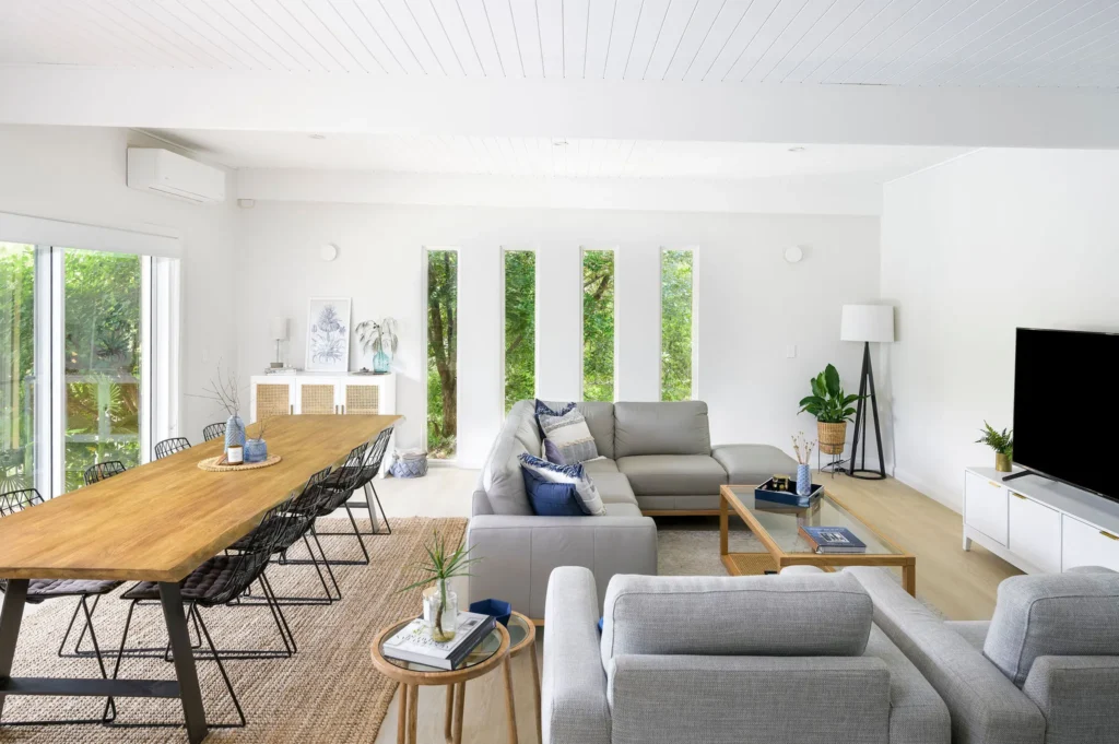 Modern living room with a cozy, light grey sectional sofa, wooden dining table, and large TV. Bright windows with lush greenery outside.
