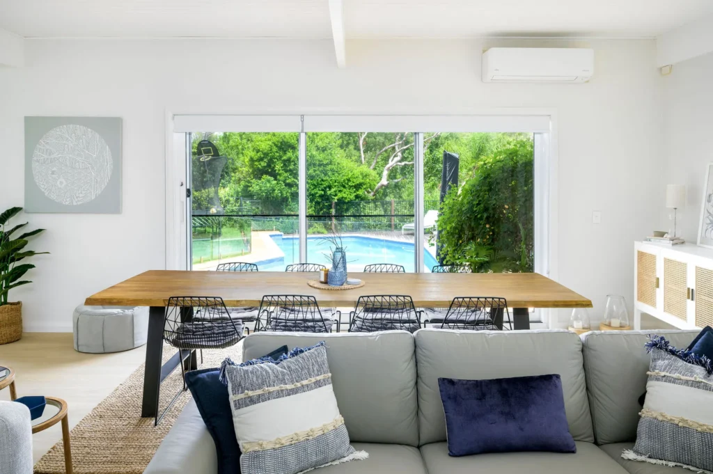 Modern living room with a gray sofa, blue and striped pillows, and a large glass door opening to a pool and lush greenery outside.