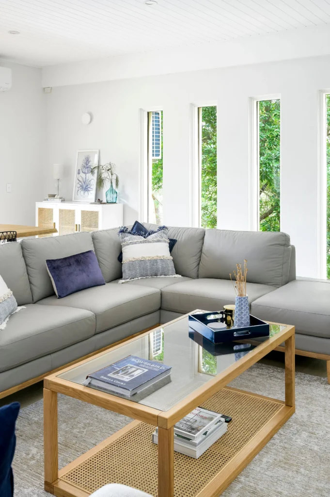 Modern living room featuring a light grey L-shaped sofa with blue and grey cushions. A wooden coffee table with a glass top holds a magazine and decor. Vertical windows reveal green foliage outside.