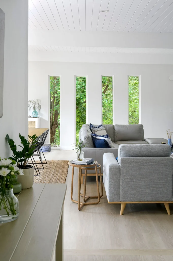 Bright and airy living room with large vertical windows revealing lush green trees. Features modern grey sofas, a wooden side table, and cozy decor.