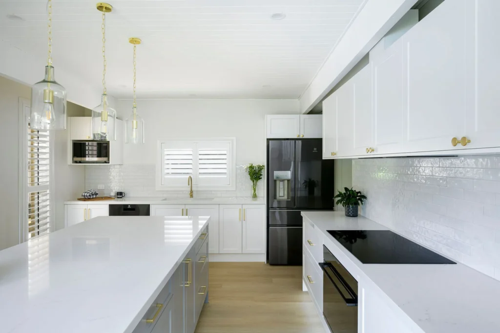Sleek modern kitchen with white cabinets, gold accents, and glass pendant lights. Features a black refrigerator, white island, and potted plants.