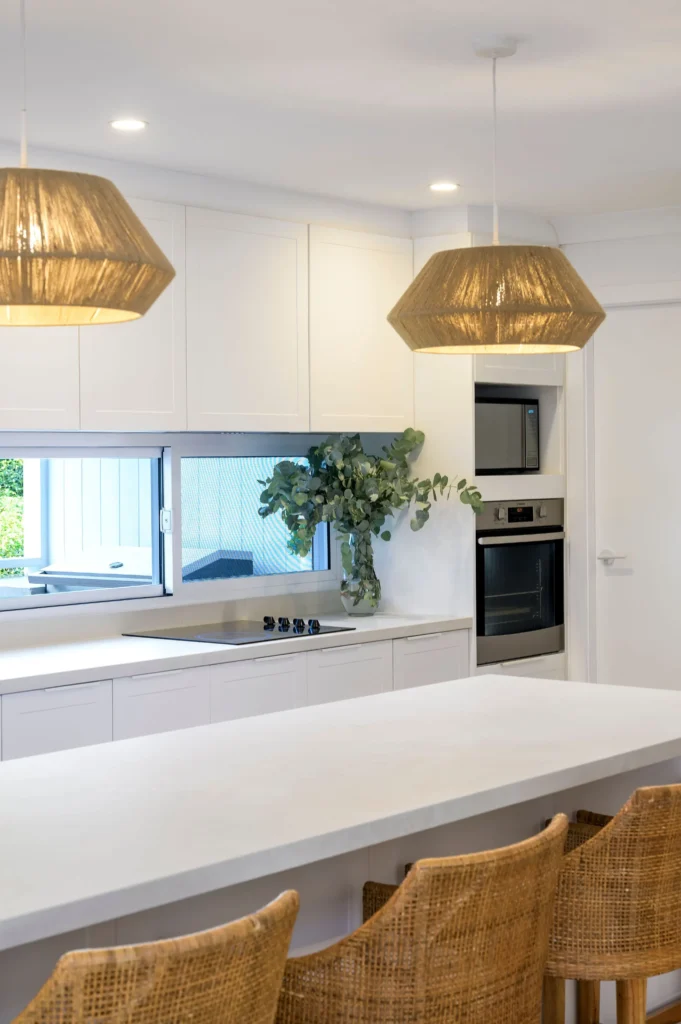 Modern white kitchen with large island, rattan bar stools, pendant lights, and greenery in a glass vase.