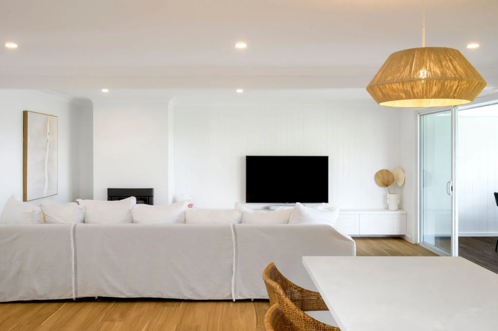 Modern living room with a cozy, light ambiance. White sofa faces a flat-screen TV on a white wall. Rattan lamp and chair add warmth. Sliding glass doors on the right.